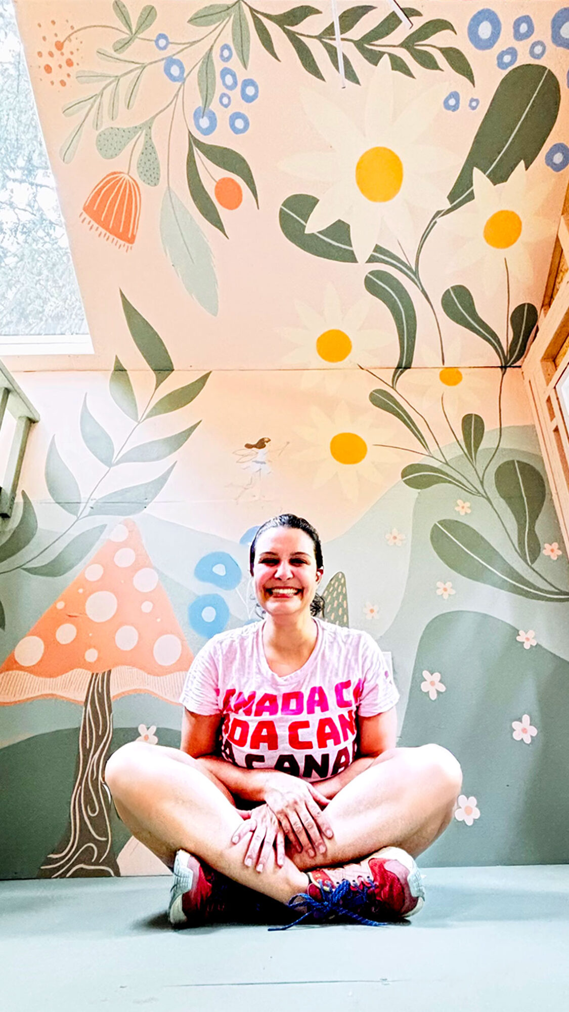 Mural artist Renata Sene sitting cross-legged and smiling in front of a completed enchanted fairy forest treehouse mural featuring mushrooms, rolling green hills, botanical leaves, white daisies, a tiny fairy, and a golden sun on a peach and sage background