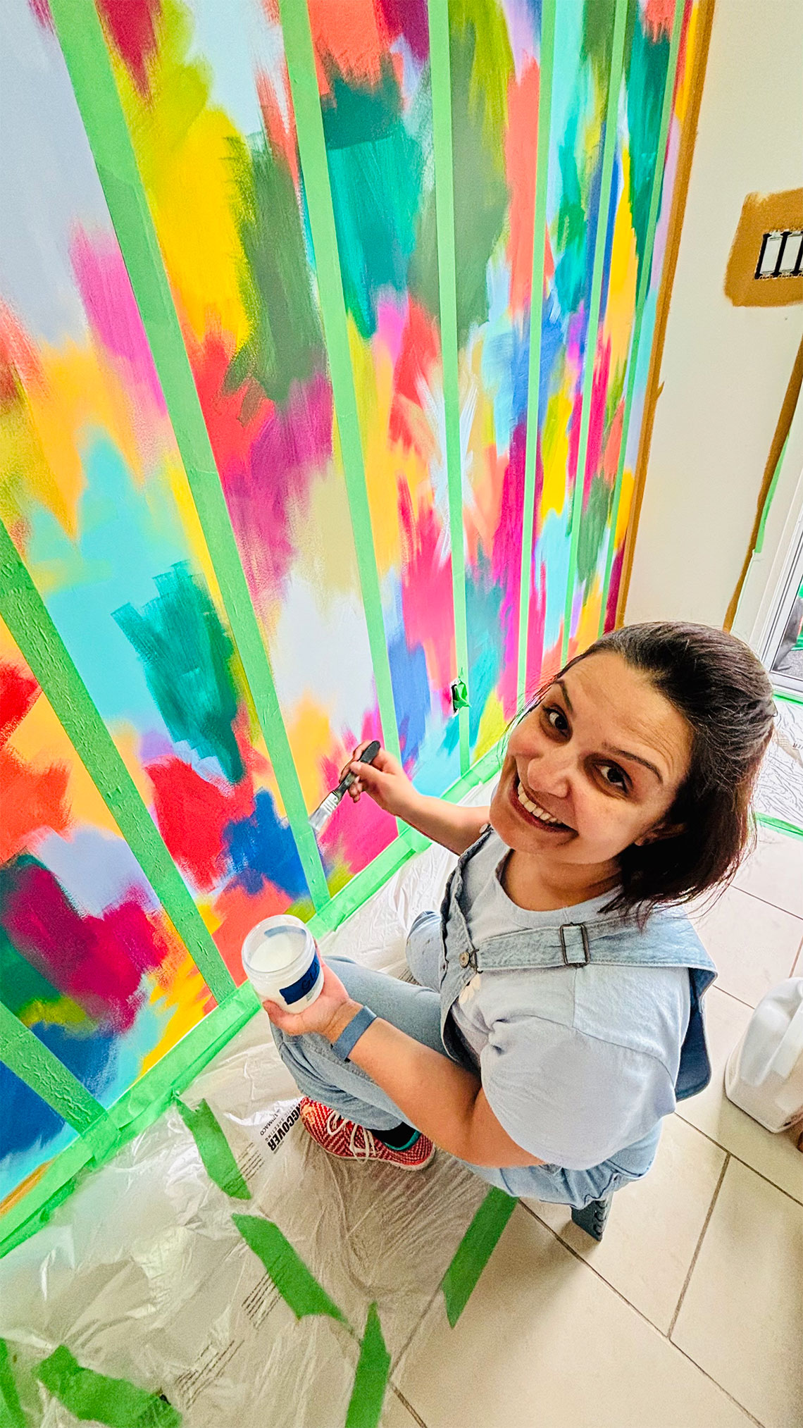 Mural artist Renata Sene smiling while painting a colourful mudroom mural with bold diagonal stripes in green, yellow, teal, pink, red, and orange using painter's tape, holding a paintbrush and paint pot, crouching on a drop-cloth covered tile floor
