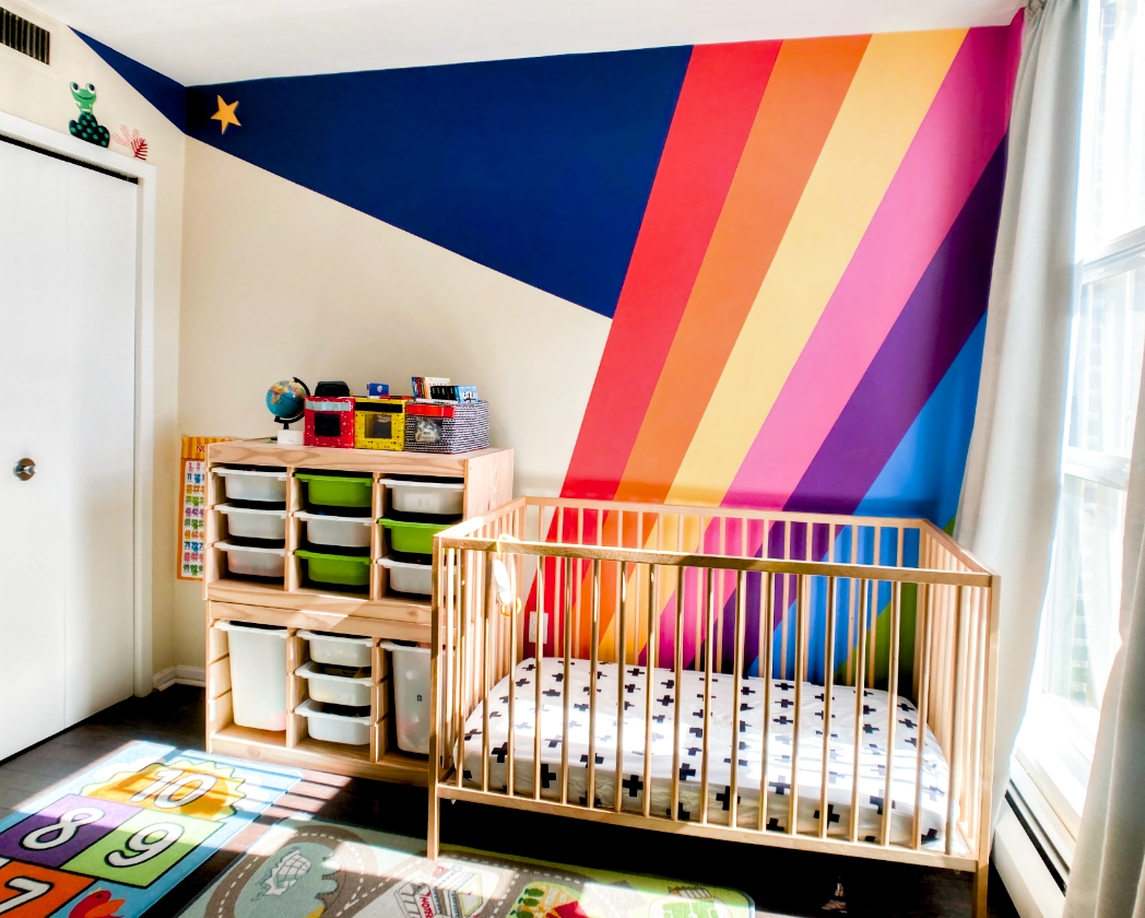Bold rainbow stripes nursery mural with navy blue geometric triangle, vibrant diagonal bands in red, orange, pink, purple, and blue behind wooden crib