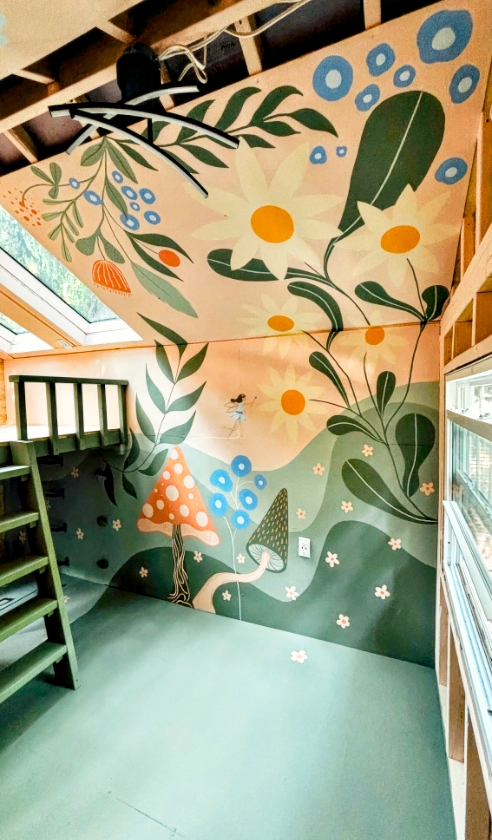 Full interior of a kids' treehouse with a hand-painted enchanted fairy forest mural covering the ceiling and walls, featuring white daisies, green leaves, blue berries, a tiny fairy, orange and grey mushrooms, and a lush green rolling landscape, with a green bunk bed ladder and windows