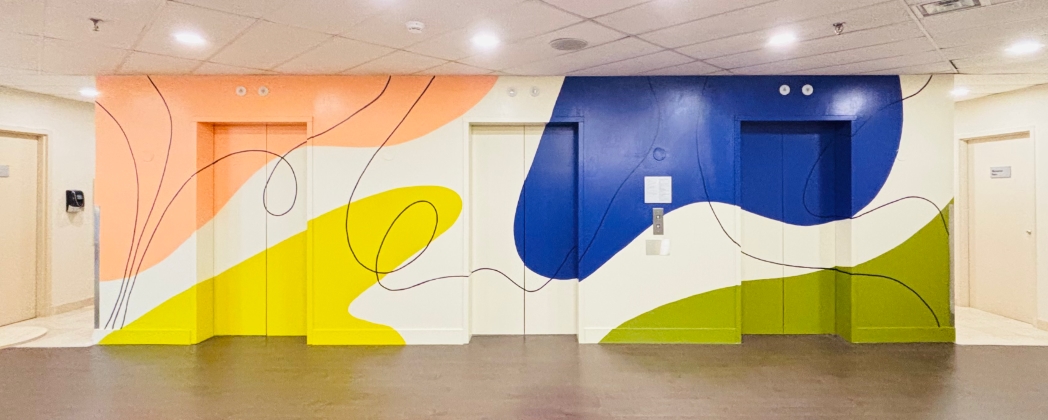 Modern abstract mural in a healthcare lobby hallway featuring large flowing organic shapes in peach, bright yellow, navy blue, and olive green with black line details on a cream background, painted across walls flanking two elevator doors
