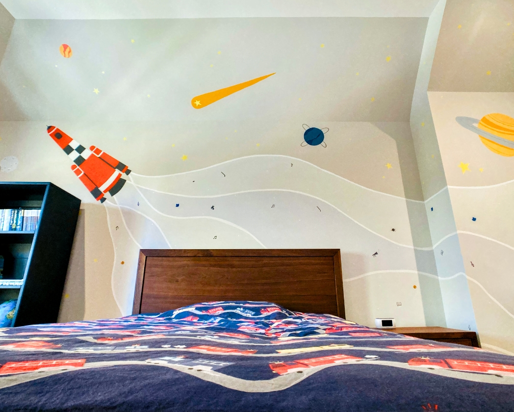 Outer space bedroom mural overhead view with orange rocket ship launching across ceiling, yellow comet, blue planet, and Saturn above kids' bed
